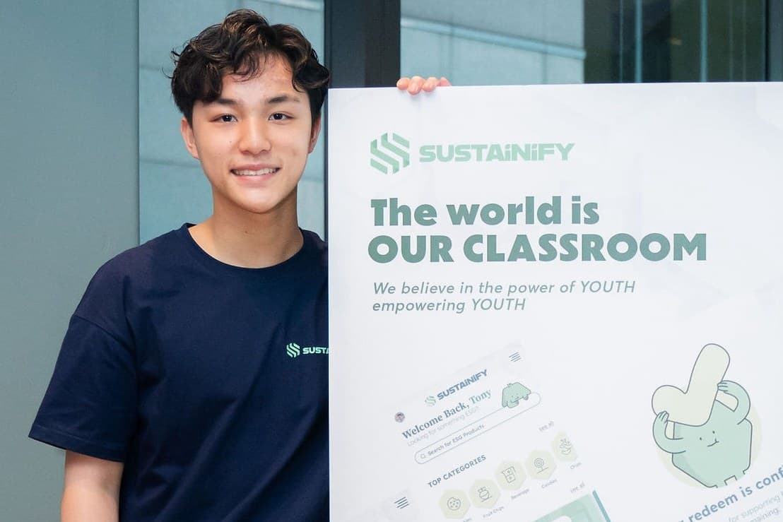 Boris Lo at the Sustainify launch, wearing a navy Sustainify T-shirt and holding a large Sustainify poster reading The world is our classroom, we believe in the power of youth empowering youth