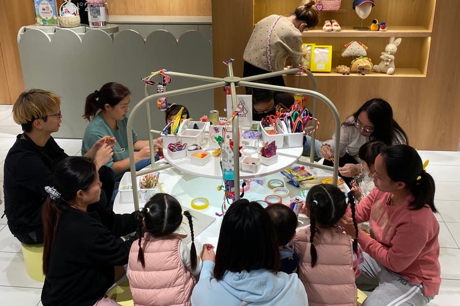 Around eight children sit on low stools at a round craft table with three adult helpers among them, sharing a central carousel of pens, tape and craft materials.