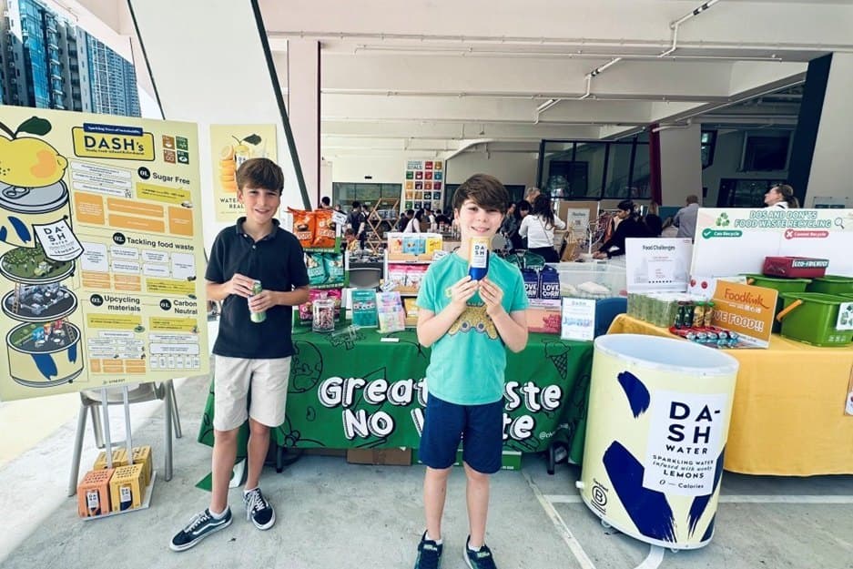 Two boys stand in a covered open-air school concourse in front of a stall with a Great Taste No Waste cloth, an illustrated board tracing a drinks can through its materials and production, and a neighbouring table of green Foodlink crates.