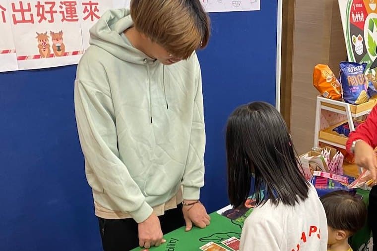 A booth with a green globe-print cloth in a decorated indoor hall, a young helper standing behind it and half a dozen children and parents leaning in over a spread of picture cards and a reusable bag.