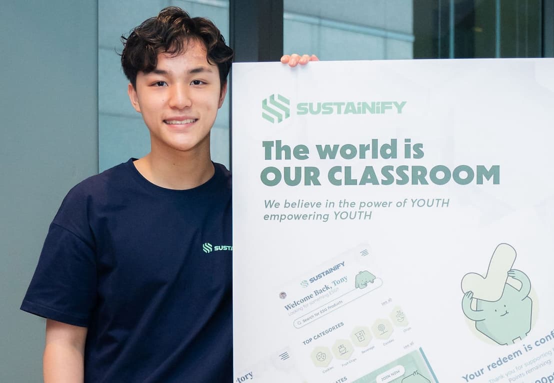 A young man in a navy Sustainify T-shirt holds up a large printed board showing screenshots of the membership platform, including a signed-in welcome screen, a row of product categories and a redemption confirmation.