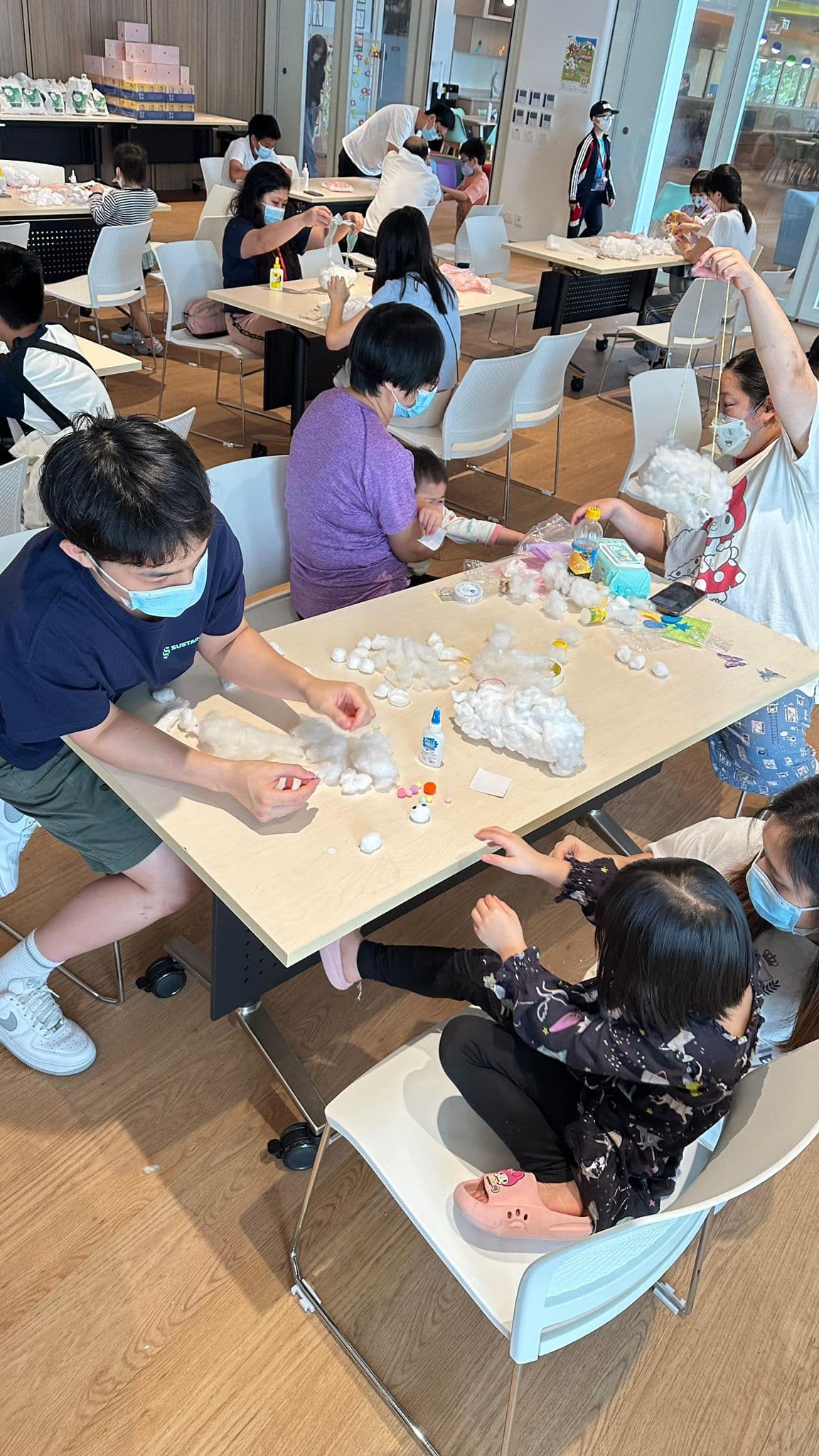 A Sustainify volunteer in a navy Sustainify T-shirt shaping cotton wool at a table alongside several children and their parents, with craft materials spread across the table