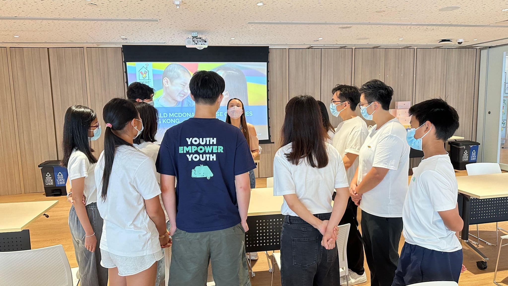 Sustainify volunteers in white T-shirts, and one in a navy Youth Empower Youth tee, standing in a semicircle while an RMHC Hong Kong staff member briefs them in front of a presentation screen