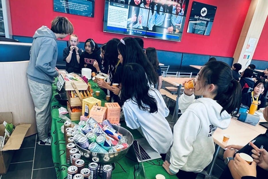 About eight students set up a long table in a school cafeteria, laying out rows of cups, cans and packets in front of a red wall with two wall-mounted screens.