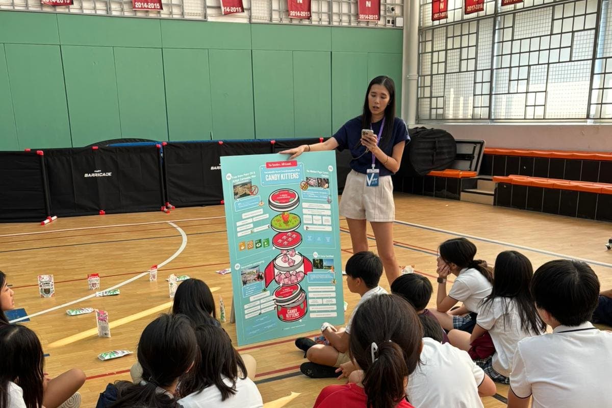 A young facilitator with a lanyard stands beside a large round information board in a school sports hall, talking to about twenty pupils sitting on the floor in front of her.