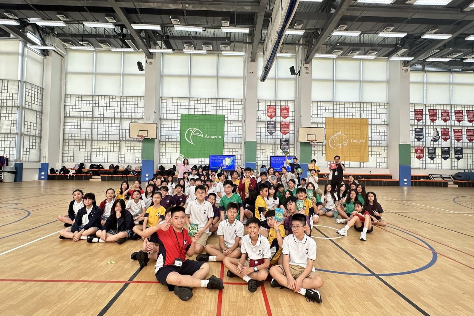 A wide view of a school sports hall with roughly a hundred pupils in coloured house shirts sitting together on the floor, with a few facilitators standing behind them.
