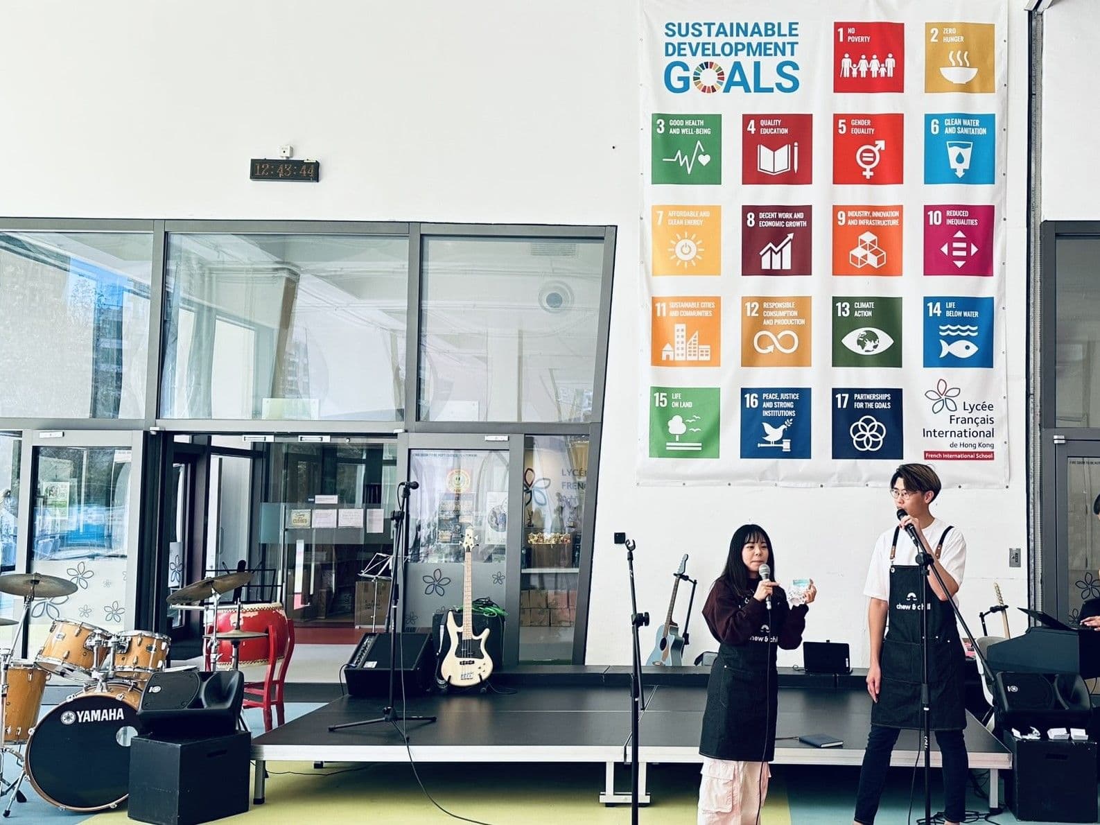 Two young people in dark aprons speak into microphones on a low stage in a school atrium, under a large wall banner of the seventeen Sustainable Development Goals, with drums and guitars behind them.