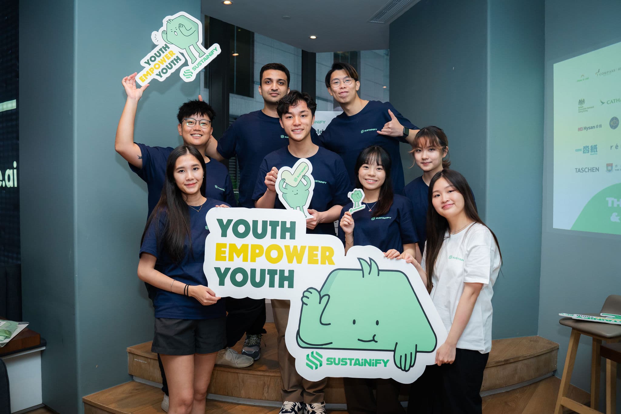 Eight Sustainify team members standing together in a bright room, holding a large "Youth Empower Youth" sign and a cut-out of the green Sustainify mascot