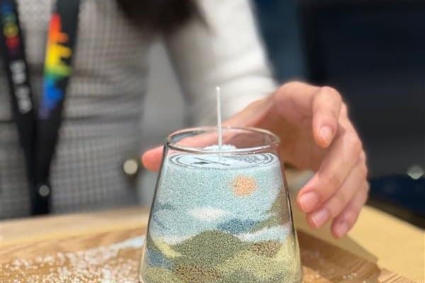 A finished glass jar of layered blue, green and sand-coloured grains sits on a wooden board, with one hand resting beside it.