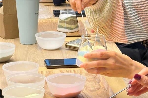 Several pairs of hands work along one table, each spooning coloured sand into their own glass jar from a row of white bowls.